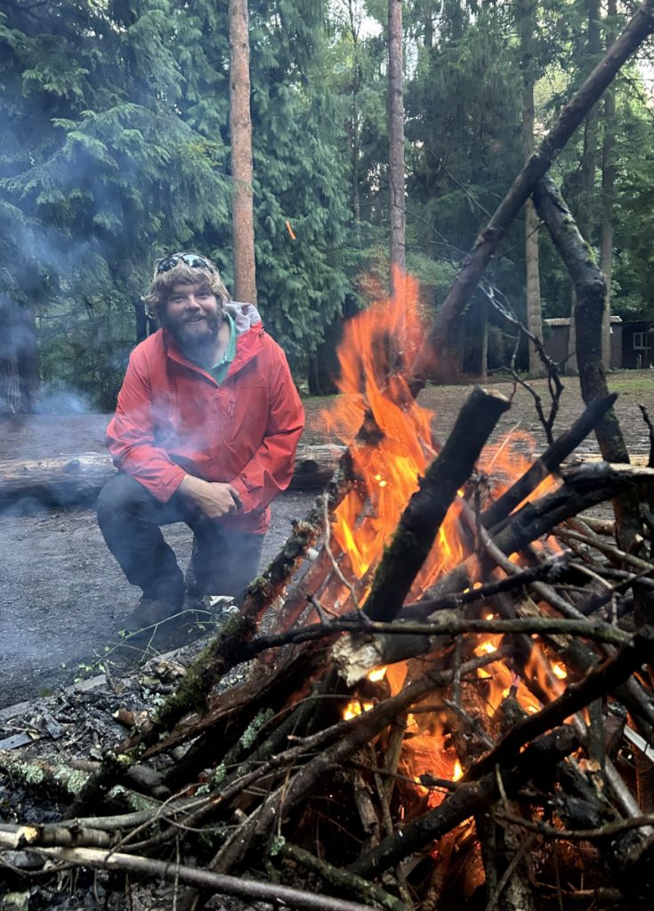 Mark kneeling by a campfire.